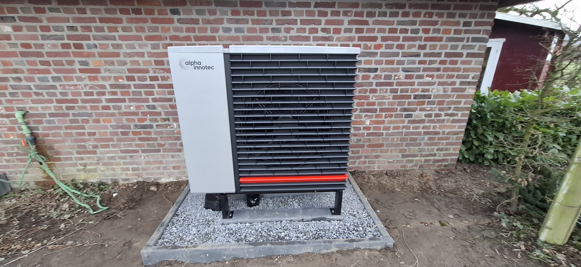 Außeneinheit einer Wärmepumpe der Marke alpha innotec auf Schotterfundament vor einer Backsteinwand installiert.